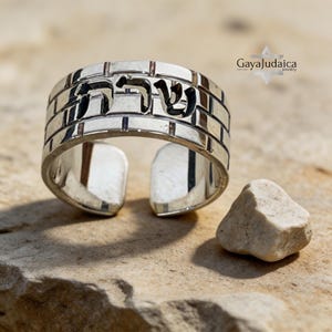 May include: A silver ring with Hebrew text, set against a brick-like pattern. The ring is open at the bottom and sits on a textured, light brown surface. A small, light-colored stone rests nearby.
