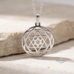 May include: A silver-toned circular pendant necklace featuring an openwork Star of David design with a single small clear stone set in the center. The pendant hangs from a delicate silver-toned chain, displaying the GayaJudaica logo in the corner.