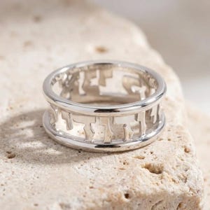 May include: A silver-toned metal ring featuring a cut-out design that incorporates Hebrew characters around the entire circumference of the band. The ring has two smooth, solid horizontal rims at the top and bottom edges, framing the openwork lettering.