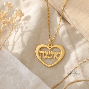 Puede incluir: Un collar dorado con un colgante en forma de corazón. El colgante presenta escritura hebrea dentro del contorno del corazón. El collar se muestra sobre una tela color crema con flores secas y una superficie de madera en el fondo.