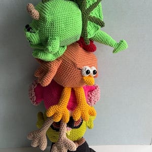 May include: A stack of four crocheted stuffed toys. The bottom toy is a black and grey penguin. The next toy is a green and yellow bird. The next toy is a pink and orange bird. The top toy is a green dinosaur with a long neck.
