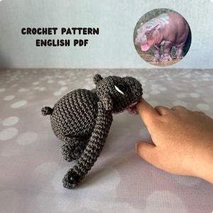 May include: A crocheted gray hippopotamus toy with black button eyes and a pink mouth. The toy is interacting with a finger. The text "CROCHET PATTERN ENGLISH PDF" is at the top. A photo of a real hippo is in the upper right corner.