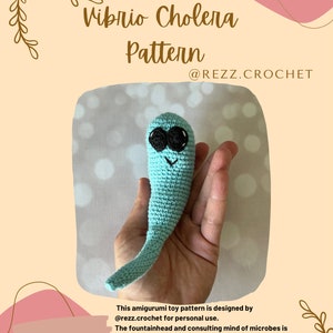 May include: A crocheted, light blue amigurumi toy in the shape of a Vibrio Cholera bacterium, with black eyes and a smiling mouth. The image includes the text "Vibrio Cholera Pattern" and the Instagram handle "@REZZ.CROCHET".