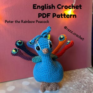 May include: A handmade blue crochet peacock with a rainbow tail. The peacock has black eyes, a tan beak, and a brown base. The text "English Crochet PDF Pattern" and "Peter the Rainbow Peacock" are displayed above the peacock. The Instagram handle "@rezz.crochet" is also visible.
