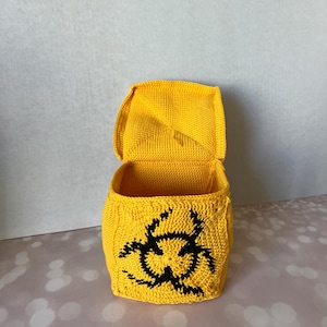May include: A yellow crocheted box with a black biohazard symbol on the front. The box is open and the inside is visible.