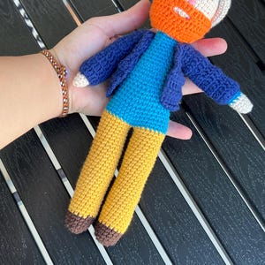 Könnte beinhalten: Eine gehäkelte Puppe mit einem weißen Hut, orangem Bart, blauer Jacke und gelber Hose. Die Puppe hält einen Pinsel.