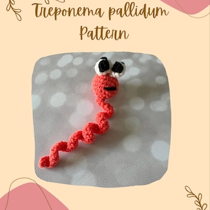 May include: A crocheted amigurumi toy shaped like a spiral bacterium with a smiling face and googly eyes. The toy is red and is on a white background with a pattern of small white circles. The text "Treponema pallidum Pattern" is above the toy.