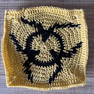 May include: A yellow crocheted square with a black design in the center. The design is a stylized swirl or flower.