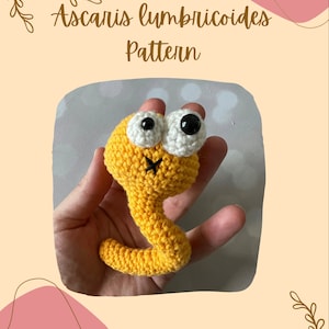 Puede incluir: Un gusano amarillo tejido a crochet con ojos blancos y una boca negra. El gusano está siendo sostenido en una mano. La imagen tiene el texto "Ascaris lumbricoides Pattern" en la parte superior.