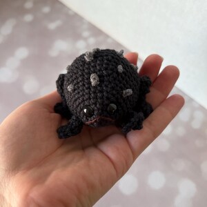 May include: A small, black crocheted frog with grey spots and black eyes. The frog is sitting in the palm of a hand.