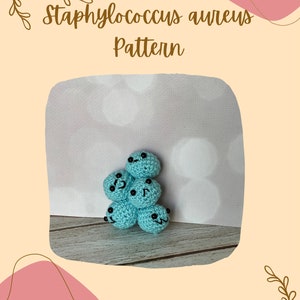 May include: A crocheted plush toy shaped like a cluster of Staphylococcus aureus bacteria. The bacteria are light blue with black eyes and smiles. The toy is on a wooden surface.