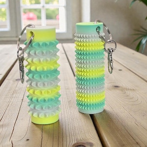 May include: Two cylindrical keychains with a textured, ridged design. The keychains are gradient in color, transitioning from yellow to green. Each keychain has a silver keyring and clasp attached.