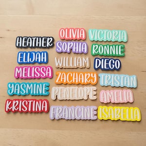 May include: A collection of colorful name tags with white lettering. Names include Heather, Elijah, Melissa, Yasmine, Kristina, Olivia, Sophia, William, Zachary, Penelope, Francine, Victoria, Ronnie, Diego, Tristan, Amelia, and Isabella.