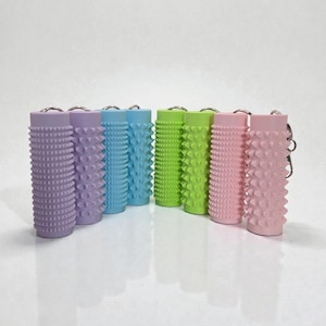 Op de afbeelding: Een verzameling kleurrijke, cilindervormige sleutelhangers. De sleutelhangers zijn in tinten lavendel, blauw, groen en roze. Elke sleutelhanger heeft een gestructureerd, noppen oppervlak en een zilverkleurige sleutelring.