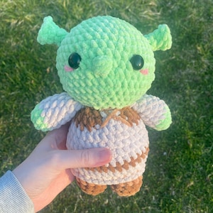 May include: A handmade green and white crochet doll with black eyes and pink cheeks. The doll has a light brown vest and feet. The doll is held in a person's hand against a background of green grass.