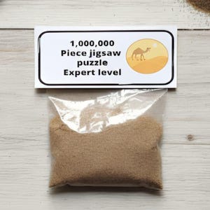 A joke "1,000,000 Piece jigsaw puzzle Expert level" gag gift. The product consists of a small, clear bag filled with fine tan sand, topped with a professional white cardstock label. The label features bold, dark text and a circular icon of a camel in a desert, set against a bright, uncluttered interior background.