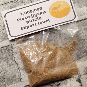 A joke "1,000,000 Piece jigsaw puzzle Expert level" gag gift. The product consists of a small, clear bag filled with fine tan sand, topped with a professional white cardstock label. The label features bold, dark text and a circular icon of a camel in a desert, set against a bright, uncluttered interior background.