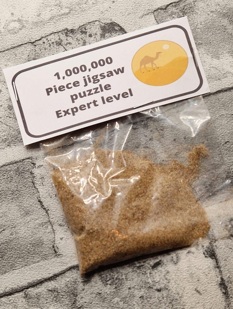 A joke "1,000,000 Piece jigsaw puzzle Expert level" gag gift. The product consists of a small, clear bag filled with fine tan sand, topped with a professional white cardstock label. The label features bold, dark text and a circular icon of a camel in a desert, set against a bright, uncluttered interior background.
