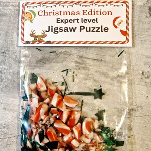 A joke "1,000,000 Piece Christmas style jigsaw puzzle Expert level" gag gift. The product consists of a small, clear bag filled with candy cane, glitter and tinsel, topped with a professional white cardstock label. The label features bold, dark text and a circular icon of a camel in a desert, set against a bright, uncluttered interior background.