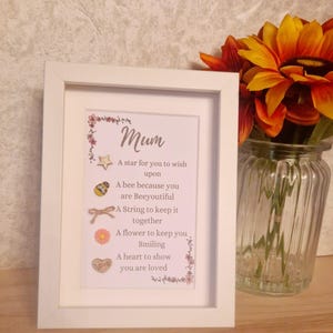 Op de afbeelding: Een wit frame met een roze en witte bloemenrand. De tekst in het frame luidt "Mum" en bevat een gedicht over een ster, een bij, een touwtje, een bloem en een hart. Het gedicht luidt: "Een ster om een wens te doen, Een bij omdat je prachtig bent, Een touwtje om alles bij elkaar te houden, Een bloem om je te laten glimlachen, Een hart om te laten zien dat je geliefd bent."
