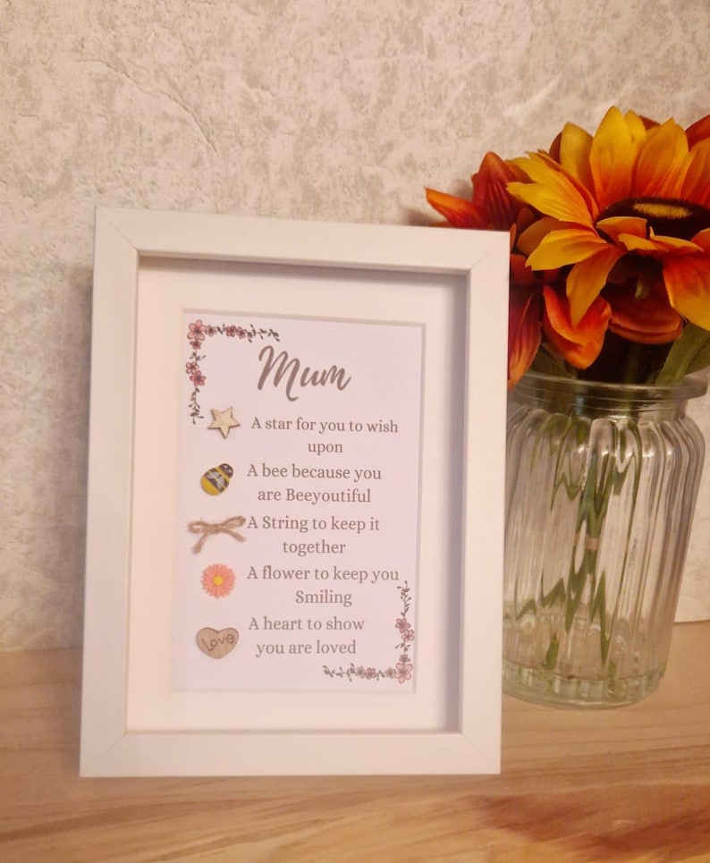 Op de afbeelding: Een wit frame met een roze en witte bloemenrand. De tekst in het frame luidt "Mum" en bevat een gedicht over een ster, een bij, een touwtje, een bloem en een hart. Het gedicht luidt: "Een ster om een wens te doen, Een bij omdat je prachtig bent, Een touwtje om alles bij elkaar te houden, Een bloem om je te laten glimlachen, Een hart om te laten zien dat je geliefd bent."