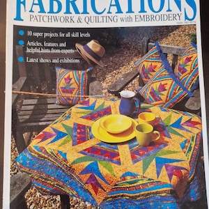 May include: A colourful patchwork quilt with a starburst pattern in shades of yellow, blue, green, and purple. The quilt is laid out on a table with a straw hat, a jug, and cups. The magazine cover reads "Fabrications Patchwork & Quilting with Embroidery".