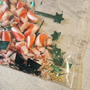 A joke "1,000,000 Piece Christmas style jigsaw puzzle Expert level" gag gift. The product consists of a small, clear bag filled with candy cane, glitter and tinsel, topped with a professional white cardstock label. The label features bold, dark text and a circular icon of a camel in a desert, set against a bright, uncluttered interior background.