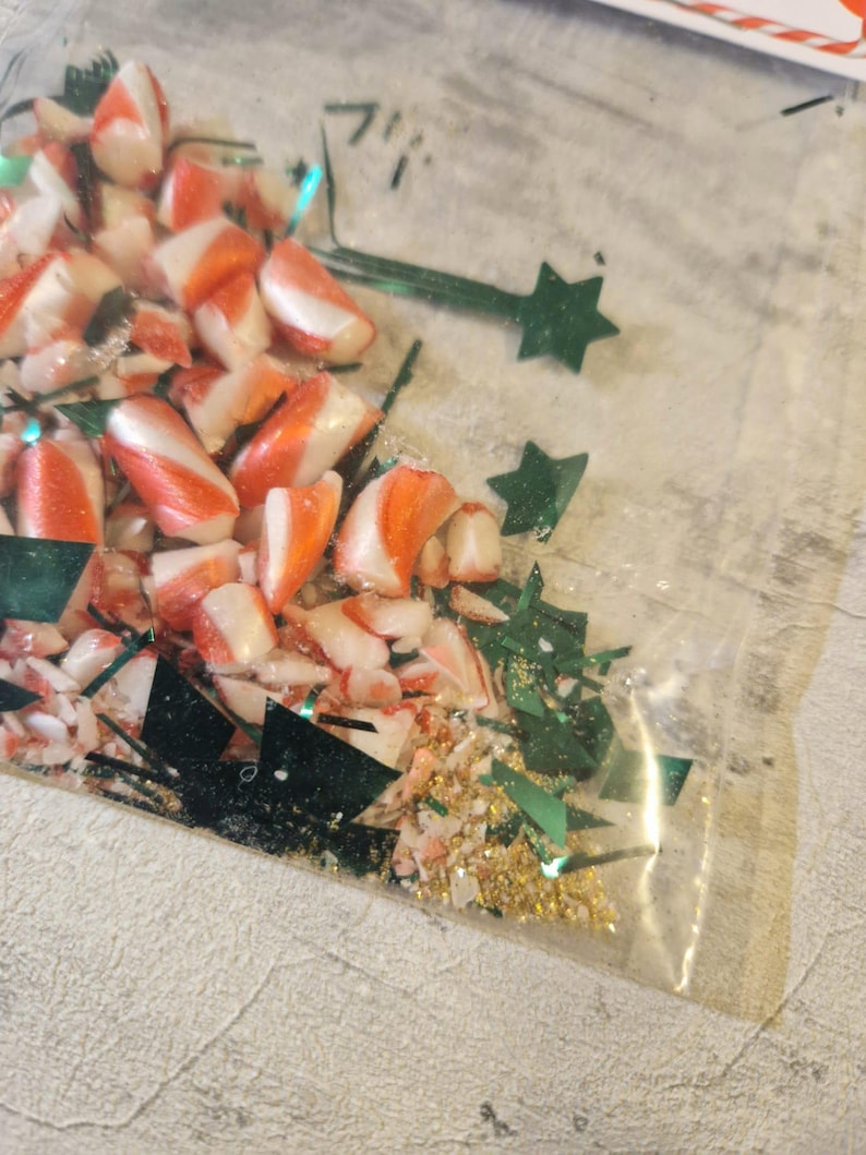 A joke "1,000,000 Piece Christmas style jigsaw puzzle Expert level" gag gift. The product consists of a small, clear bag filled with candy cane, glitter and tinsel, topped with a professional white cardstock label. The label features bold, dark text and a circular icon of a camel in a desert, set against a bright, uncluttered interior background.