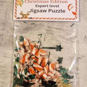 A joke "1,000,000 Piece Christmas style jigsaw puzzle Expert level" gag gift. The product consists of a small, clear bag filled with candy cane, glitter and tinsel, topped with a professional white cardstock label. The label features bold, dark text and a circular icon of a camel in a desert, set against a bright, uncluttered interior background.