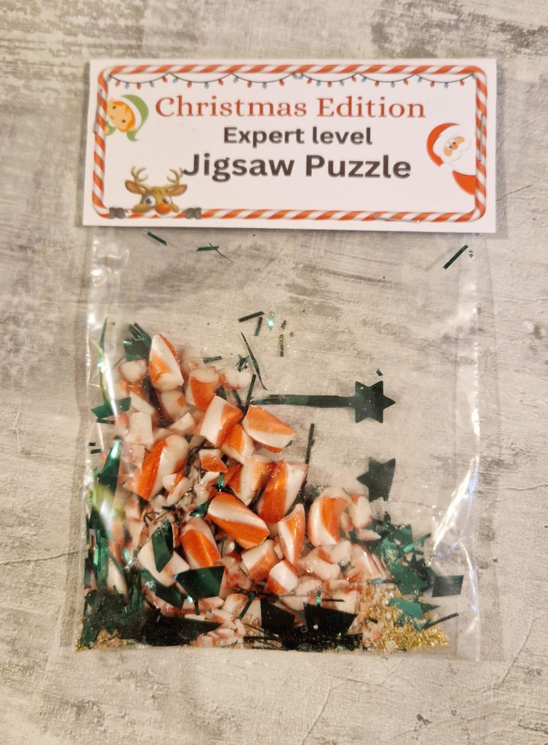 A joke "1,000,000 Piece Christmas style jigsaw puzzle Expert level" gag gift. The product consists of a small, clear bag filled with candy cane, glitter and tinsel, topped with a professional white cardstock label. The label features bold, dark text and a circular icon of a camel in a desert, set against a bright, uncluttered interior background.