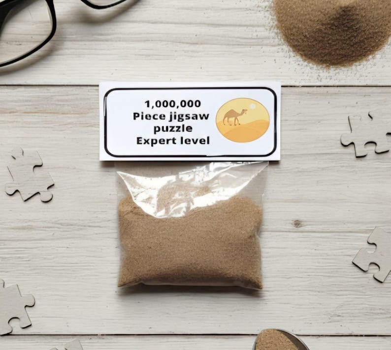 A joke "1,000,000 Piece jigsaw puzzle Expert level" gag gift. The product consists of a small, clear bag filled with fine tan sand, topped with a professional white cardstock label. The label features bold, dark text and a circular icon of a camel in a desert, set against a bright, uncluttered interior background.