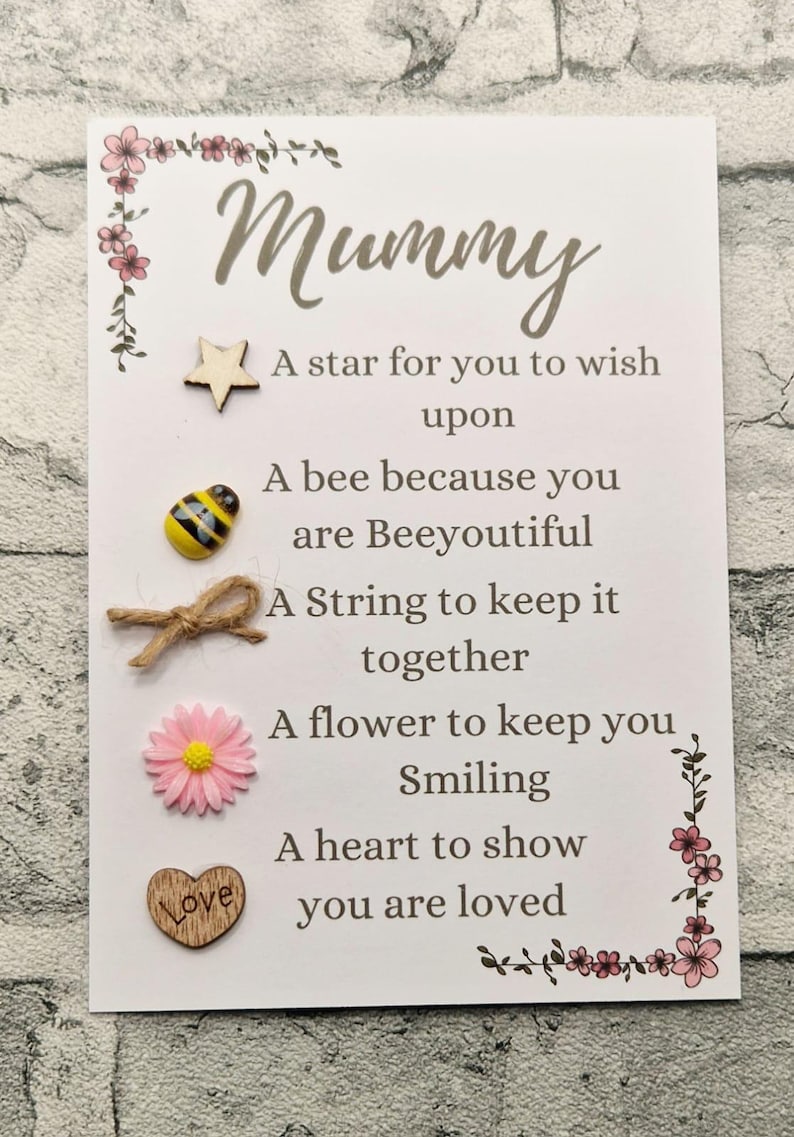 Op de afbeelding: Een witte kaart met het woord "Mummy" geschreven in bruin cursief. De kaart heeft een gedicht met de volgende tekst: "Een ster om een wens te doen, Een bij omdat je prachtig bent, Een touwtje om alles bij elkaar te houden, Een bloemetje om je te laten glimlachen, Een hart om te laten zien dat je geliefd bent". De kaart heeft een kleine houten ster, een klein houten hart met het woord "Love" erop, een kleine houten bij, een kleine roze bloem en een klein stukje touw.