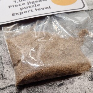 A joke "1,000,000 Piece jigsaw puzzle Expert level" gag gift. The product consists of a small, clear bag filled with fine tan sand, topped with a professional white cardstock label. The label features bold, dark text and a circular icon of a camel in a desert, set against a bright, uncluttered interior background.