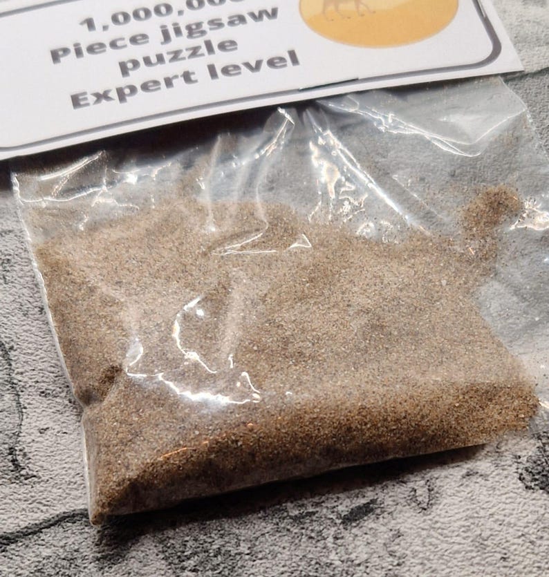 A joke "1,000,000 Piece jigsaw puzzle Expert level" gag gift. The product consists of a small, clear bag filled with fine tan sand, topped with a professional white cardstock label. The label features bold, dark text and a circular icon of a camel in a desert, set against a bright, uncluttered interior background.