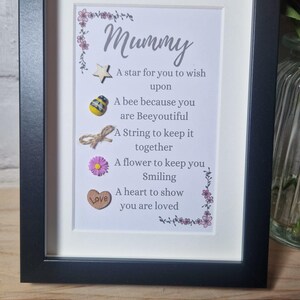 Op de afbeelding: Een ingelijst afdruk met een witte achtergrond en een zwarte lijst. De afdruk bevat een gedicht over een moeder met de woorden "Mummy" en "A star for you to wish upon" en andere zinnen. Het gedicht is versierd met kleine houten en stoffen versieringen.