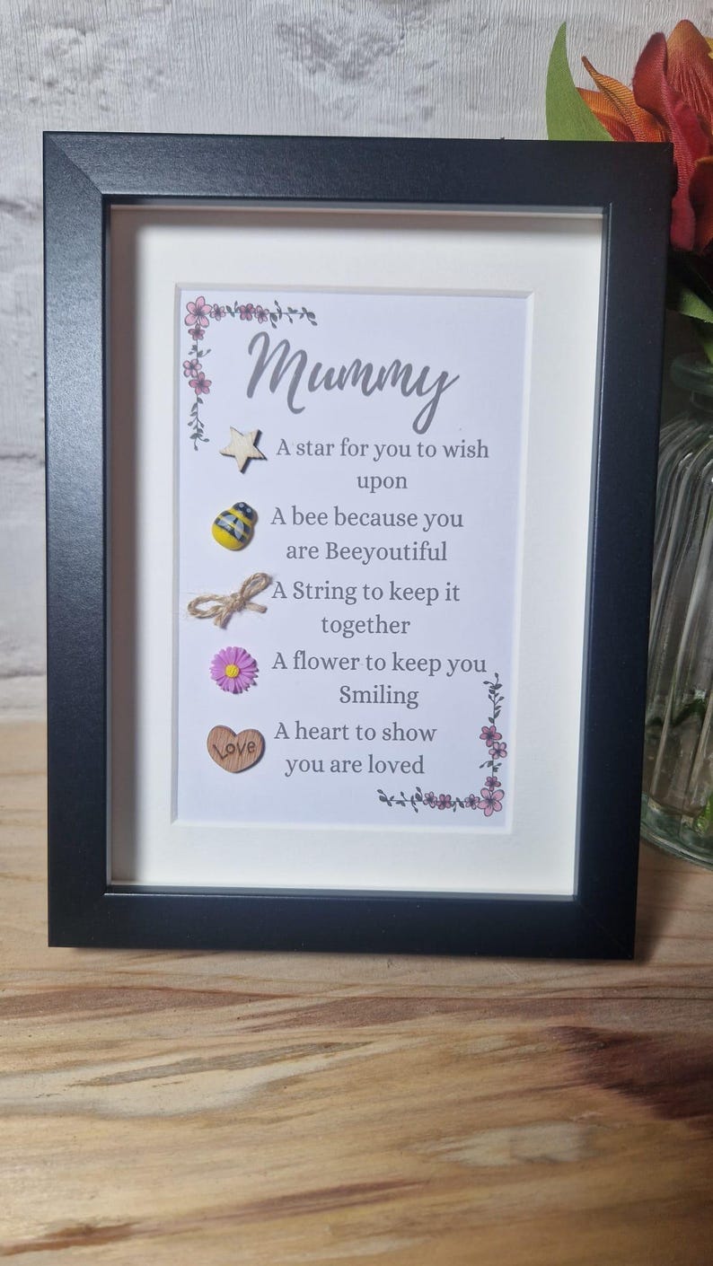 Op de afbeelding: Een ingelijst afdruk met een witte achtergrond en een zwarte lijst. De afdruk bevat een gedicht over een moeder met de woorden "Mummy" en "A star for you to wish upon" en andere zinnen. Het gedicht is versierd met kleine houten en stoffen versieringen.
