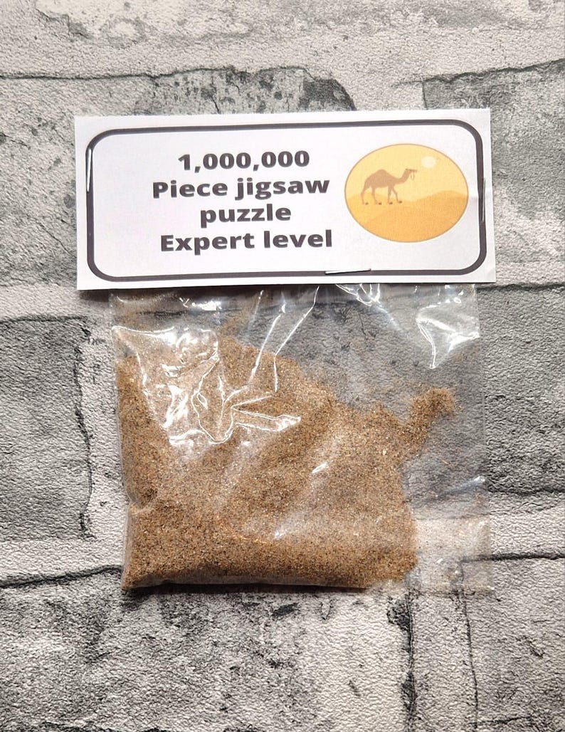 A joke "1,000,000 Piece jigsaw puzzle Expert level" gag gift. The product consists of a small, clear bag filled with fine tan sand, topped with a professional white cardstock label. The label features bold, dark text and a circular icon of a camel in a desert, set against a bright, uncluttered interior background.