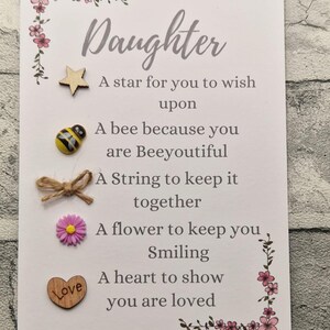 Op de afbeelding: Een witte kaart met een roze bloemrand en de tekst "Daughter" in een cursief schrift. De kaart heeft een ster, een bij, een touwtje, een bloem en een hart met het woord "Love" erop. De kaart heeft ook de tekst "A star for you to wish upon", "A bee because you are Beeyoutiful", "A String to keep it together", "A flower to keep you Smiling", "A heart to show you are loved".