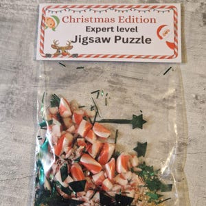 A joke "1,000,000 Piece Christmas style jigsaw puzzle Expert level" gag gift. The product consists of a small, clear bag filled with candy cane, glitter and tinsel, topped with a professional white cardstock label. The label features bold, dark text and a circular icon of a camel in a desert, set against a bright, uncluttered interior background.