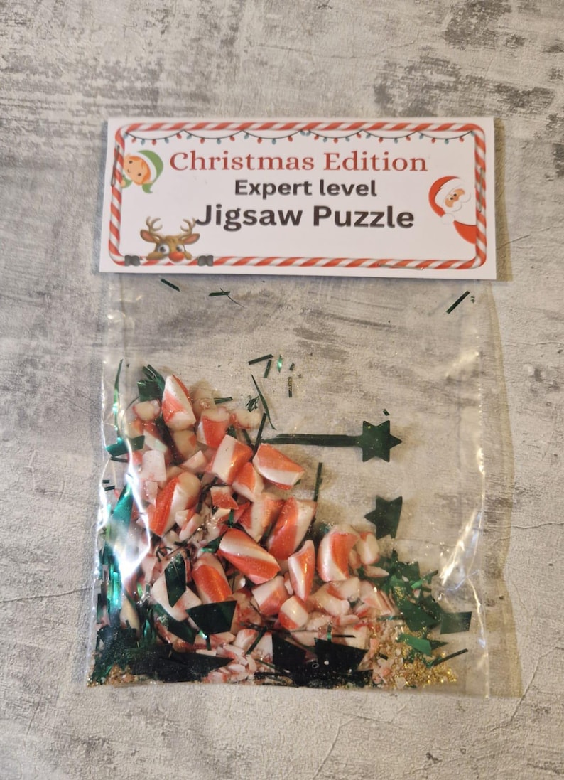 A joke "1,000,000 Piece Christmas style jigsaw puzzle Expert level" gag gift. The product consists of a small, clear bag filled with candy cane, glitter and tinsel, topped with a professional white cardstock label. The label features bold, dark text and a circular icon of a camel in a desert, set against a bright, uncluttered interior background.