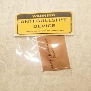 May include: A yellow warning sign with black text that reads "WARNING ANTI BULLSH*T DEVICE Please see reverse for instructions". The sign is in a clear plastic bag with a brown paper sticker that reads "ANTI BULLSHIT DEVICE" in black marker.