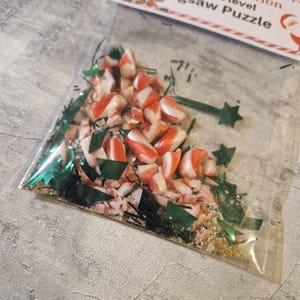 A joke "1,000,000 Piece Christmas style jigsaw puzzle Expert level" gag gift. The product consists of a small, clear bag filled with candy cane, glitter and tinsel, topped with a professional white cardstock label. The label features bold, dark text and a circular icon of a camel in a desert, set against a bright, uncluttered interior background.