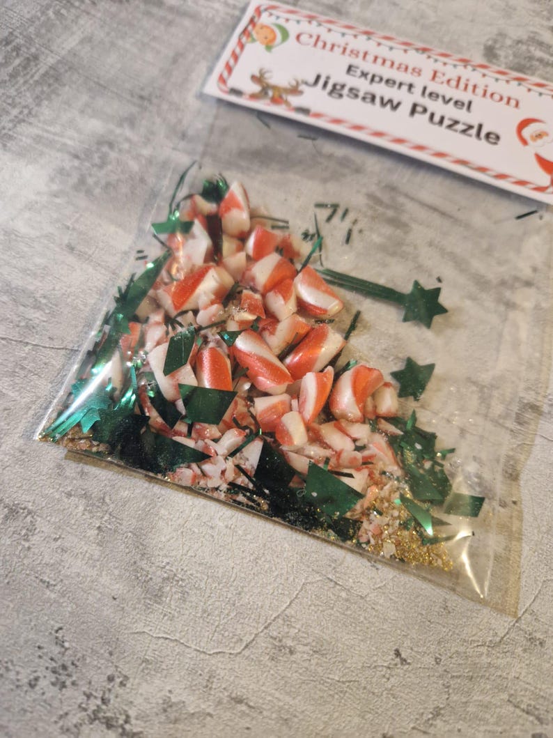 A joke "1,000,000 Piece Christmas style jigsaw puzzle Expert level" gag gift. The product consists of a small, clear bag filled with candy cane, glitter and tinsel, topped with a professional white cardstock label. The label features bold, dark text and a circular icon of a camel in a desert, set against a bright, uncluttered interior background.