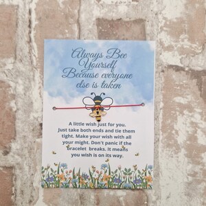 May include: A red string bracelet with a gold bee charm. The card attached to the bracelet reads "Always Bee Yourself Because everyone else is taken. A little wish just for you. Just take both ends and tie them tight. Make your wish with all your might. Don't panic if the bracelet breaks. It means you wish is on its way."