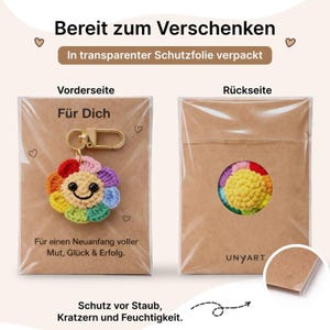 Puede incluir: Un llavero de flor arco&iacute;ris de ganchillo con un cierre dorado, empaquetado en una funda de pl&aacute;stico transparente. La parte delantera del paquete dice "F&uuml;r Dich" y "F&uuml;r einen Neuanfang voller Mut, Gl&uuml;ck & Erfolg." La parte trasera muestra el reverso de la flor.