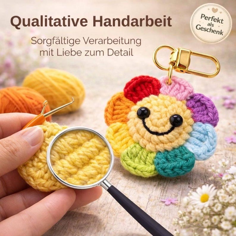 Puede incluir: Un llavero de flores de ganchillo de colores, con una cara sonriente. El llavero tiene un cierre dorado y est&aacute; hecho de varios colores de hilo. La imagen tambi&eacute;n muestra texto: "Qualitative Handarbeit" y "Perfekt als Geschenk".