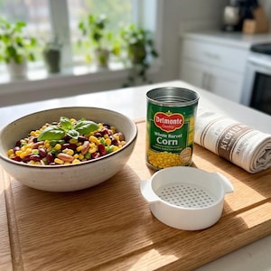 May include: A wooden cutting board with a bowl of mixed beans and corn, garnished with basil. A can of Del Monte whole kernel corn, a white strainer, and a rolled kitchen towel are also on the board. The scene is set in a bright kitchen.