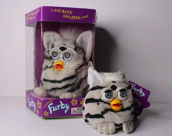 May include: Two white and black striped Furby toys. The larger Furby is in a purple and white box with the text "I will keep amazing you!" The smaller Furby has a purple tag that says "Furby" with a white star.