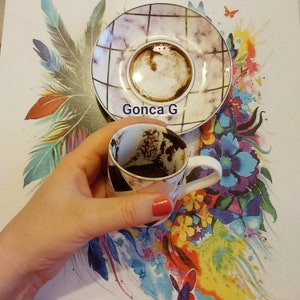 K&ouml;nnte beinhalten: Eine wei&szlig;e Kaffeetasse mit einer dunkelbraunen Fl&uuml;ssigkeit und Kaffeesatz darin. Die Tasse steht auf einer bunten, floral gemusterten Oberfl&auml;che. Der Text "Gonca G" ist auf der Oberfl&auml;che sichtbar.