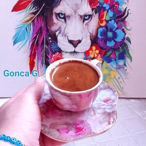 K&ouml;nnte beinhalten: Eine Nahaufnahme einer rosa-wei&szlig;en Kaffeetasse mit floralem Muster und dunkelbraunem Kaffee darin. Die Tasse steht auf einer passenden Untertasse. Die Tasse wird von einer Hand gehalten, die ein blaues Armband mit einem goldenen Stern-Anh&auml;nger tr&auml;gt. Der Hintergrund ist ein Gem&auml;lde eines L&ouml;wen mit einer bunten Blumenkrone.